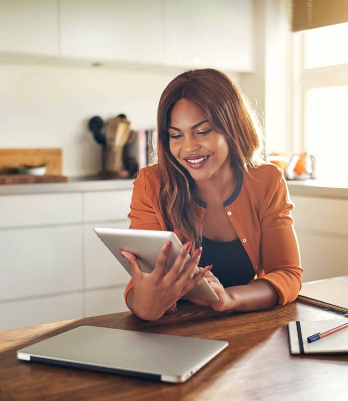 Smiling young female entrepreneur using a tablet in her kitchen Smiling young African female entrepreneur working online with a digital tablet while sitting at a table in her kitchen