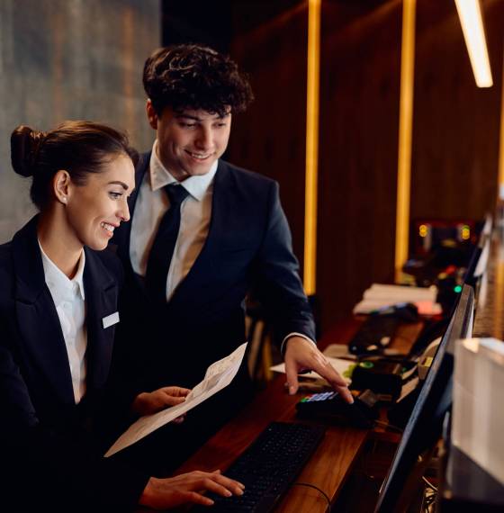Happy receptionists cooperating while working on computer at hotel front desk. Happy hotel managers through reservation list on desktop PC at reception desk.